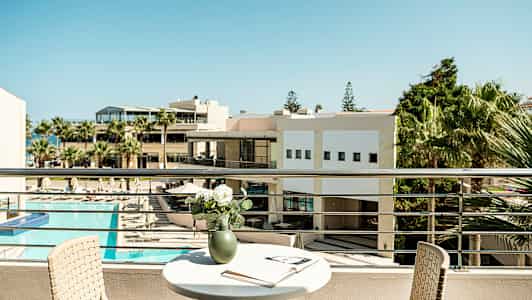 Udsigt fra en altan med et bord, en stol og en vase med blomster, samt en åben bog. I baggrunden ses en bygning og solområde med pool. (AI-genereret tekst)