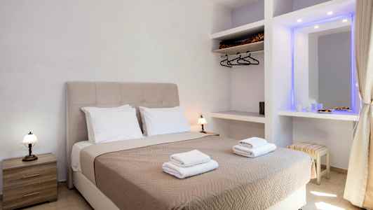 Et hotelværelse med en dobbeltseng udstyret med beige sengetæppe og to sæt håndklæder, samt sidebord og garderobe. (AI-genereret tekst)