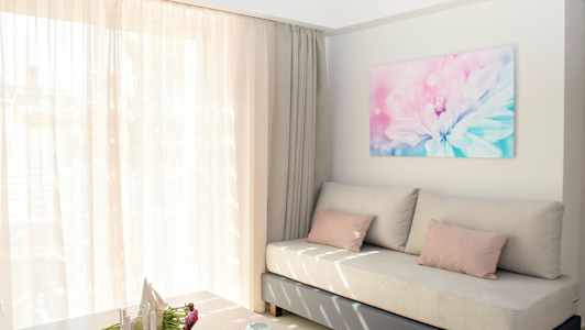 En lys og luftig stue med en beige sofa, rosa puder og et stort vindue med tynde gardiner. På væggen hænger et blomsterbillede. (AI-genereret tekst)