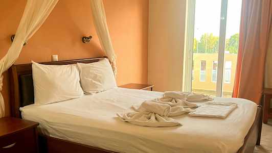 Et soveværelse med en dobbeltseng, hvide lagener og en orange væg. Et stort vindue med gardiner er synligt i baggrunden. (AI-genereret tekst)