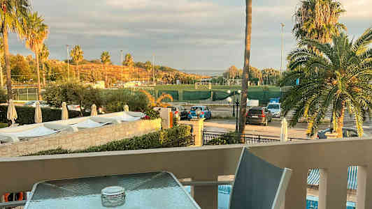 Udsigt fra en terrasse mod en sportsplads omgivet af palmer og parkeringspladser. (AI-genereret tekst)