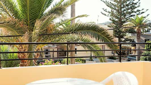 Udsigt fra altan med et bord og plastikstol, palmetræer og bygninger i baggrunden. (AI-genereret tekst)