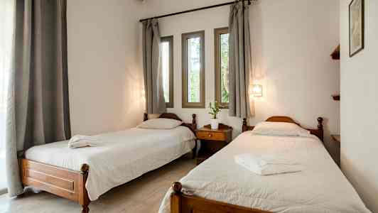 Et værelse med to enkeltsenge, et natbord imellem, hvide sengelinned og grå gardiner over vinduerne. (AI-genereret tekst)