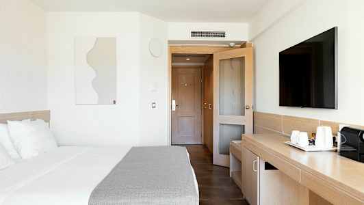 Et moderne hotelværelse med en seng, et skrivebord, et fjernsyn og en dør i baggrunden. (AI-genereret tekst)