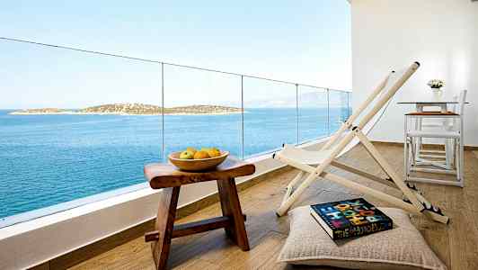 Udsigt fra en altan med stole, et bord og en skål med frugt på et træbord med havet og øer i baggrunden. (AI-genereret tekst)