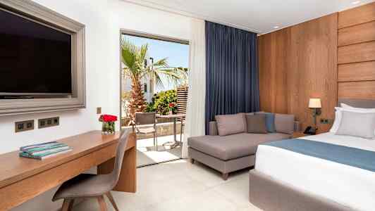 Et hotelværelse med en seng, en sofa, et skrivebord og et stort TV. Udsigt til en terrasse med træer og møbler. (AI-genereret tekst)