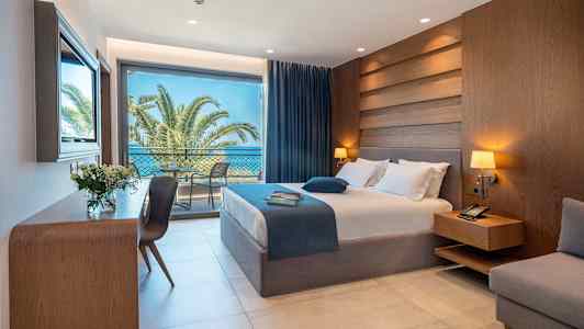 Et hotelværelse med seng, natbord, skrivebord og store glasdøre, der fører til en balkon med udsigt over palmer og havet. (AI-genereret tekst)