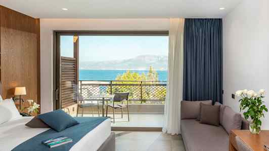 Hotelværelse med seng, sofa, skrivebord og udsigt over havet fra en balkon. (AI-genereret tekst)