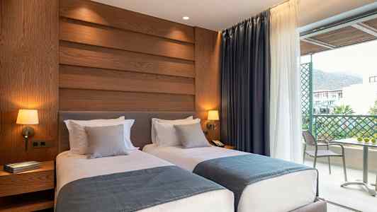 Hotelværelse med to enkeltsenge, dekoreret med blå sengetæpper og grå puder, væglamper samt udsigt til en altan. (AI-genereret tekst)