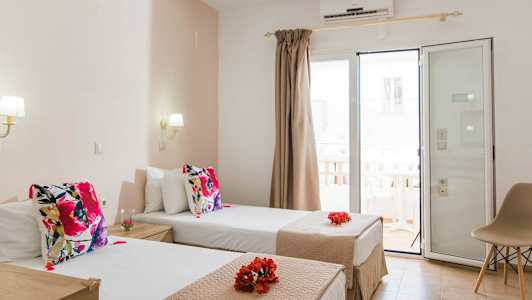 Hotelværelse med to enkeltsenge dekoreret med farverige puder og blomster. Glasdør med gardiner fører til altan. (AI-genereret tekst)