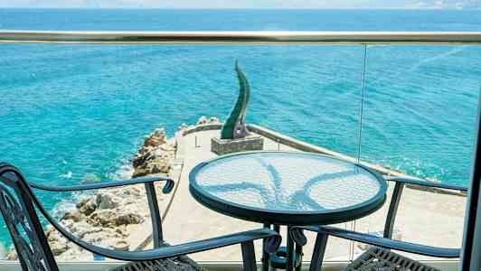 Udsigt fra en terrasse mod havet med to stole og et bord i forgrunden. (AI-genereret tekst)