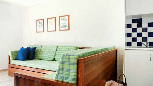 En træsofa med grønne ternede hynder, to blå puder og tre indrammede billeder på væggen. En del af et køkken med ternede fliser ses. (AI-genereret tekst)