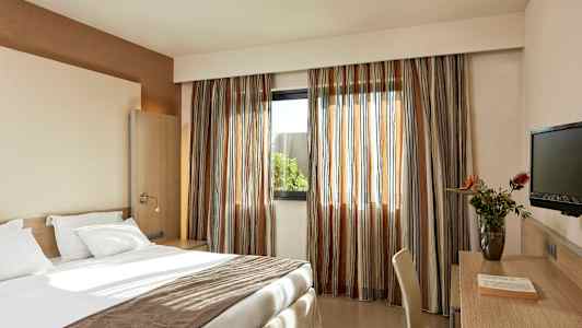 Et hotelværelse med en redt seng, skrivebord med stol, en TV og et vindue med gardiner. (AI-genereret tekst)