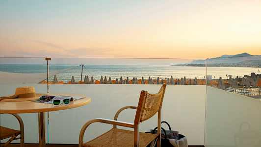 Udsigt fra en terrasse mod havet med solnedgang og strandparasoller i baggrunden. (AI-genereret tekst)