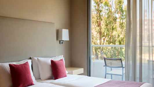 Hotelværelse med to senge, røde puder og udsigt til en altan med stole og træer udenfor. (AI-genereret tekst)