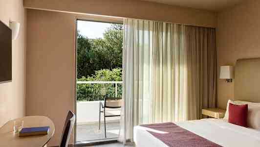 Et hotelværelse med en dobbeltseng og et skriveområde ved siden af. Værelset har et stort vindue, der fører ud til en altan med en stol og udsigt over grønne områder. (AI-genereret tekst)