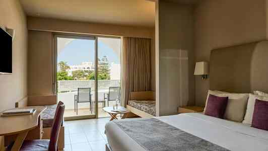 Et moderne hotelværelse med en dobbeltseng, skrivebord, siddepladser og et stort vindue, der fører ud til en balkon med udsigt over en have. (AI-genereret tekst)