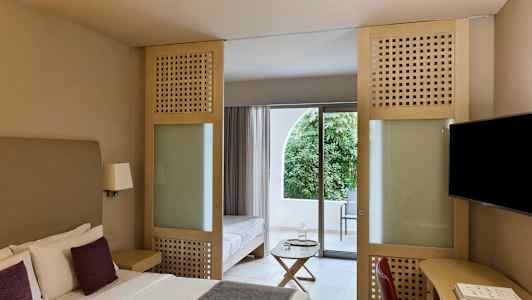 Hotelværelse med dobbeltseng, siddepladser og glasdør, der fører til en balkon med udsigt over grønt område. (AI-genereret tekst)