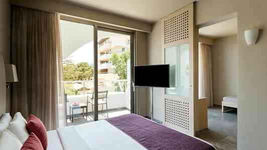 Et hotelværelse med en seng, et TV, en altan med stole og bord samt udsigt over grønne områder og en bygning. (AI-genereret tekst)