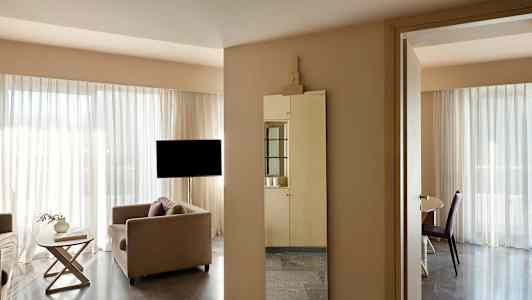 En moderne stue med store vinduer og lyse gardiner. En sofa, et lille bord og et spejl er synlige i rummet. (AI-genereret tekst)