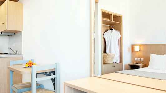 Et lyst hotelværelse med et lille køkkenområde, et bord med to stole og en seng. En hvid kjole hænger i skabet. (AI-genereret tekst)