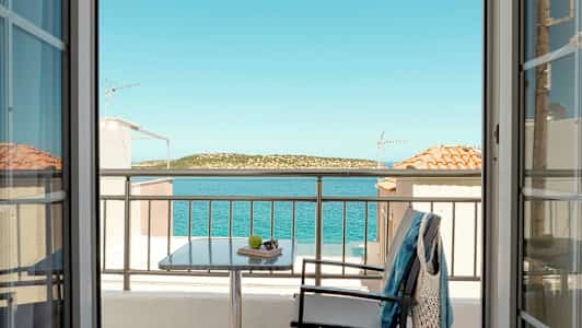 Udsigt fra et værelse gennem døre mod en balkon med bord, stol og tøj, samt hav og landskab i baggrunden. (AI-genereret tekst)