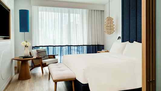 Et moderne hotelværelse med en stor seng, en lænestol, et bord og blomster samt stilrene gardiner og belysningsdetaljer. (AI-genereret tekst)