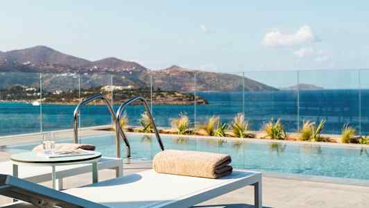 En udendørs pool med udsigt over havet og bjergene, omgivet af glasrækværk og liggestole. (AI-genereret tekst)