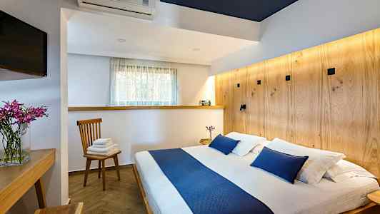 Billede af et soveværelse med en dobbeltseng og blå detaljer, et træbord, en stol med håndklæder og et tv. (AI-genereret tekst)