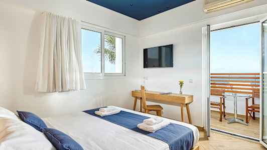 Hotelværelse med seng beklædt med blåt og hvidt sengetøj, skrivebord med stol, TV på væggen og balkon med udsigt over havet. (AI-genereret tekst)