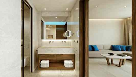 Et hotelværelse med et åbent badeværelse, der har et moderne design med en håndvask i sten og håndklæder opstillet i hylder. I baggrunden er en lys sofagruppe med blå puder og et sofabord. (AI-genereret tekst)