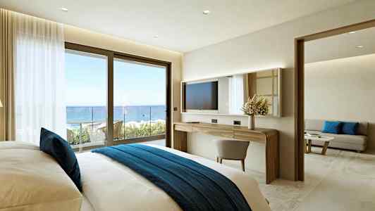 Et hotelværelse med en komfortabel seng med et blåt sengetæppe, et skrivebord med spejl og en skydedør, der fører til en balkon med havudsigt. (AI-genereret tekst)