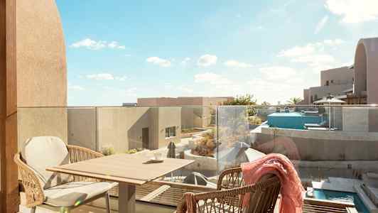 Udsigt fra en terrasse med glasrækværk. To kurvestole står ved et lille bord. Udsigt over moderne bygninger, en pool og terrasseområder under en klar blå himmel. (AI-genereret tekst)