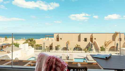En udsigt over en resort med pool og hav i det fjerne. I forgrunden ses en terrasse med en stol og et bord. (AI-genereret tekst)