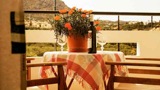 En terrasse med et bord dækket med en ternet dug, en potteplante, en vinflaske og to glas. Baggrunden viser en naturskøn udsigt. (AI-genereret tekst)