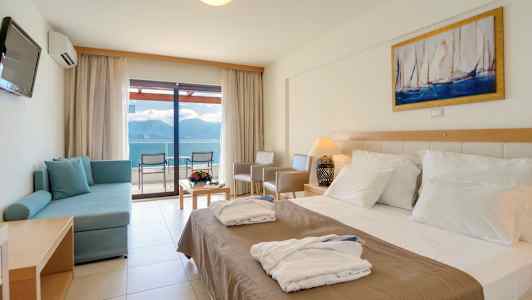 Et hotelværelse med en dobbeltseng, en sofa, et bord med blomster og udsigt til havet gennem glasdøre. (AI-genereret tekst)