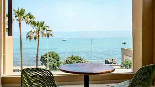 Udsigt fra et værelse mod havet, med palmer og stole på en terrasse. (AI-genereret tekst)