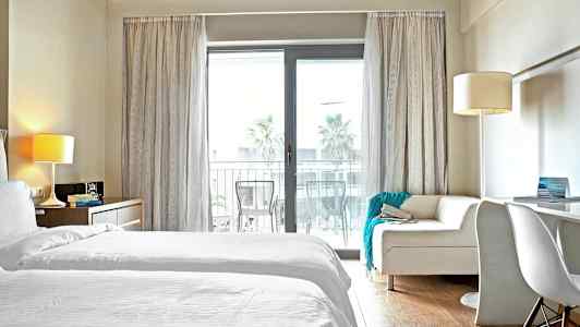 Et hotelværelse med hvidt sengetøj og en sofa foran en glasdør til en altan. (AI-genereret tekst)