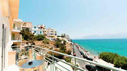 Udsigt fra en altan mod havet med biler parkeret langs en vej og bygninger i nærheden. (AI-genereret tekst)