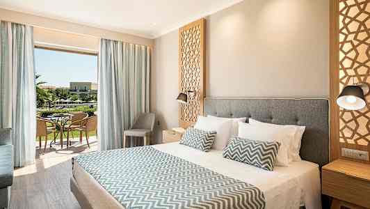 Et hotelværelse med en stor seng, dekorative puder og en åbning mod en altan med udsigt over haven. (AI-genereret tekst)