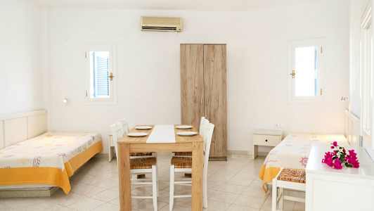 Et lyst værelse med to enkeltsenge, et spisebord med stole og et garderobeskab. (AI-genereret tekst)