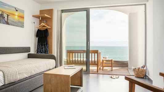 Et hotelværelse med havudsigt. En seng og et sofabord foran en glasdør, der fører ud til en veranda. (AI-genereret tekst)