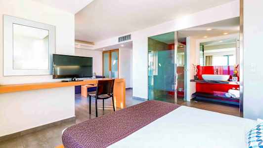 Et moderne hotelværelse med en seng i forgrunden, tv, skrivebord og et åbent badeværelsesdesign med en rød accentvæg. (AI-genereret tekst)