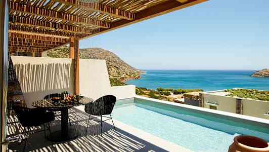 Udsigt over en terrasse med pool, bord og stole samt havet og bjergene i baggrunden. (AI-genereret tekst)