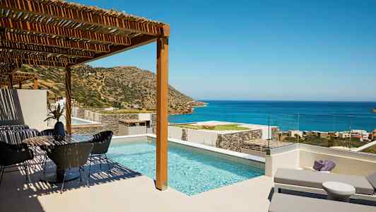 Billede af en terrasse med pool, solsenge og spiseområde, med udsigt over havet og bjergene under en klar himmel. (AI-genereret tekst)