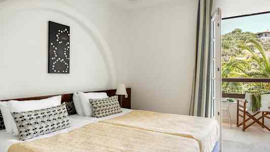 Værelset viser to enkeltsenge med guldfarvede sengetæpper og en dekorativ pude på hver seng. En dekoration hænger over hovedgærdet, og et vindue med udsigt til grønt og et hus ses til højre. (AI-genereret tekst)