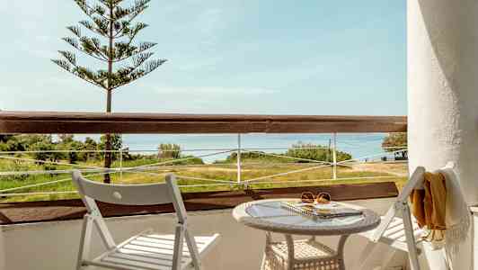 En balkon med udsigt over landskab og hav, med to hvide stole og et bord. (AI-genereret tekst)