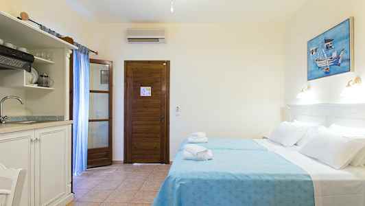 Et hotelværelse med to senge beklædt med blå sengelinned, en træbeklædt dør, et lille køkken med vask og et maleri på væggen. (AI-genereret tekst)