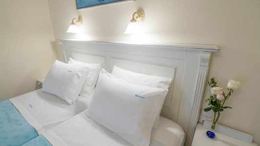 En opredt dobbeltseng med hvide lagner, et blåt sengetæppe, hvide puder, et billede ovenover og en lille vase med blomster på et natbord. (AI-genereret tekst)