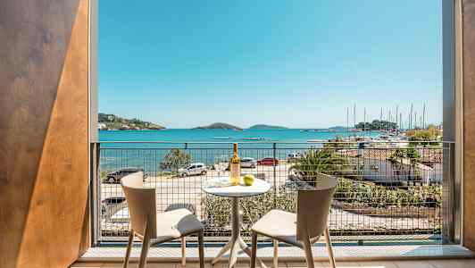 Udsigt fra en balkon med to stole og et bord, mod havet og en lystbådehavn. (AI-genereret tekst)